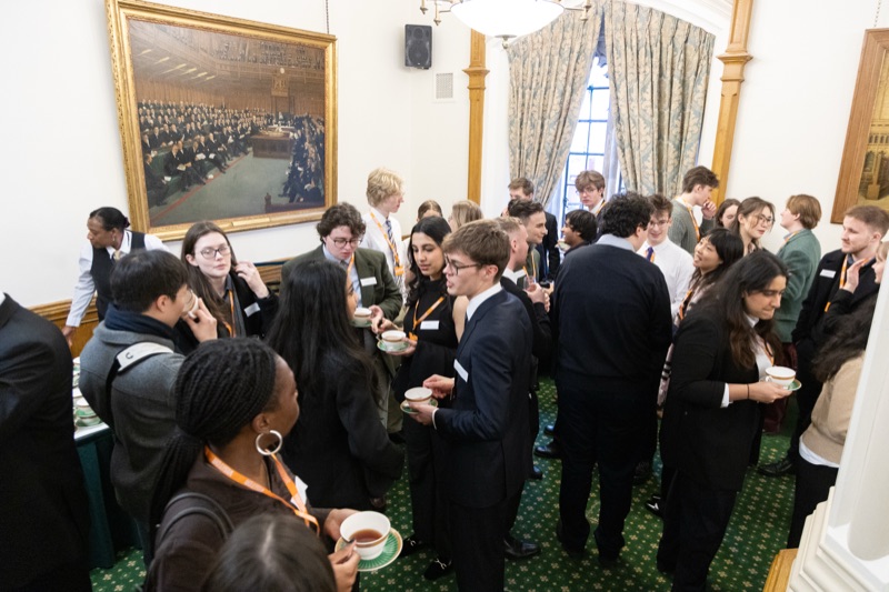 Attendees holding teacups beneath a large painting of the House of Commons chamber