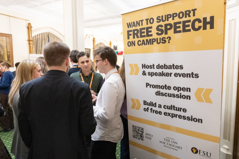 Attendees in conversation beside a banner reading Want to support free speech on campus? Host debates and speaker events, promote open discussion, build a culture of free expression