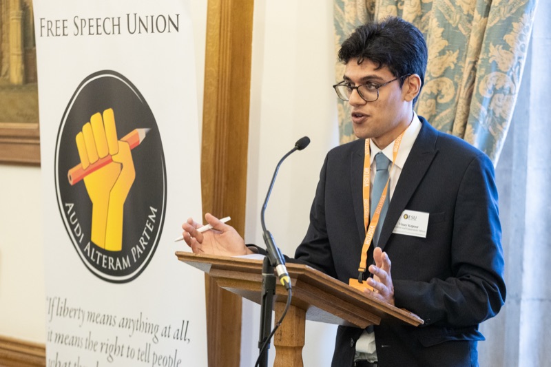 A student speaking from a lectern beside the Free Speech Union banner, which reads Audi Alteram Partem